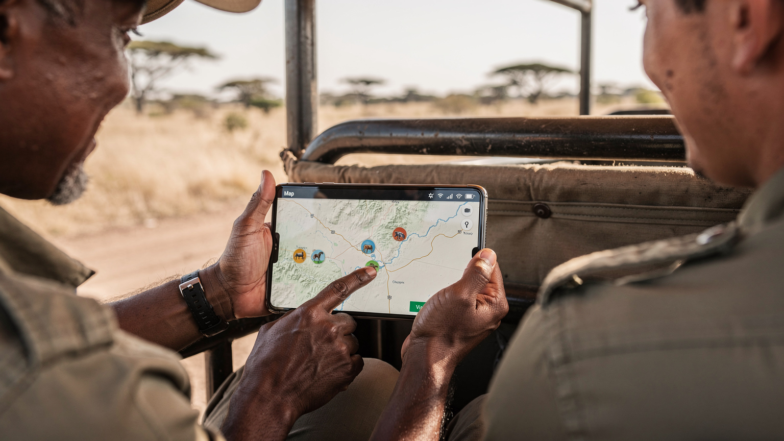 Safari průvodce v terénním vozidle ukazuje turistovi na obrazovku telefonu mapu oblasti s vyznačenými zvířaty.