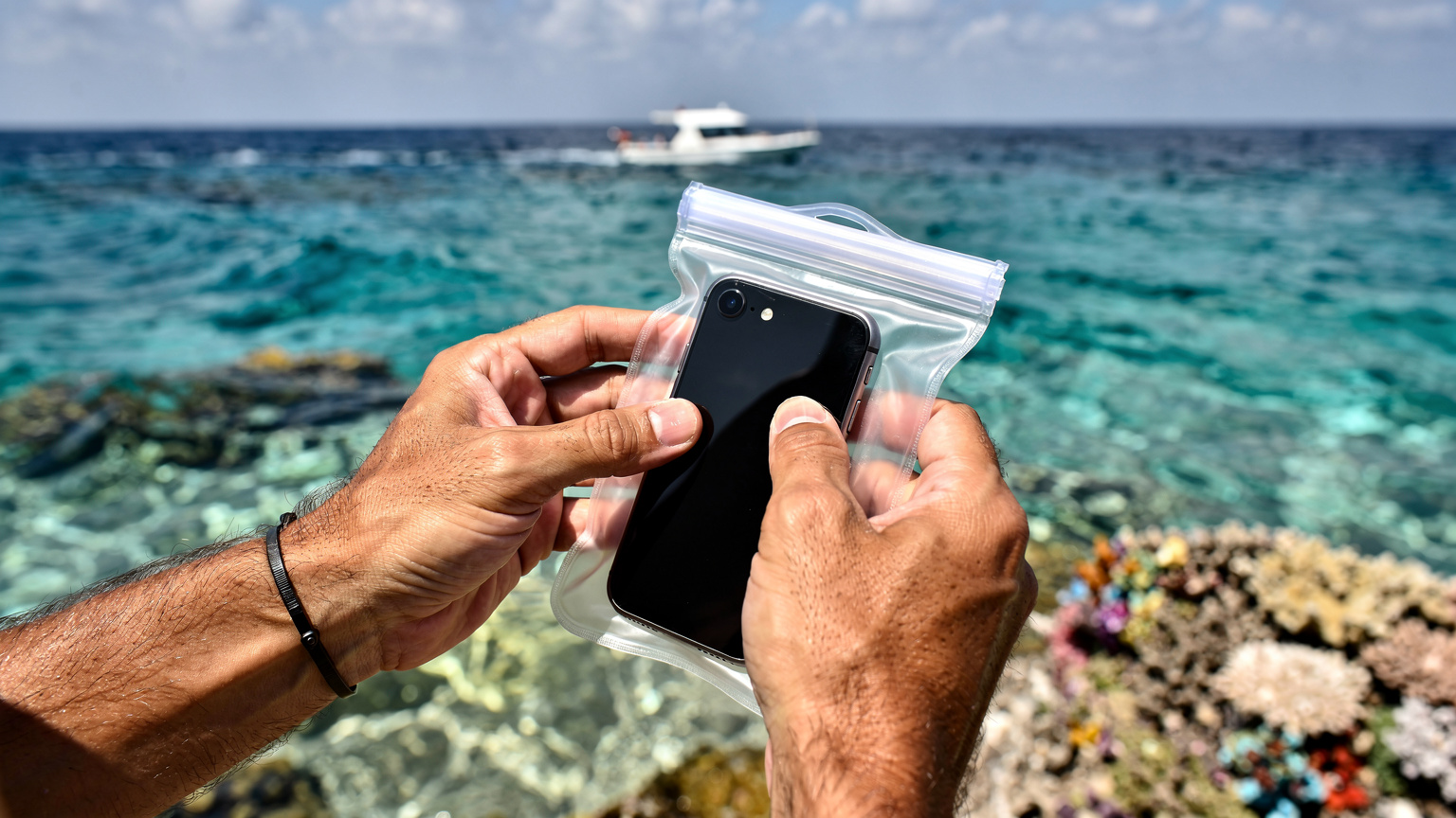 Un uomo sta immergendo il suo telefono in una custodia impermeabile trasparente, preparandosi per un'attività di snorkeling nelle acque cristalline del Mar Rosso, con una barca in lontananza.