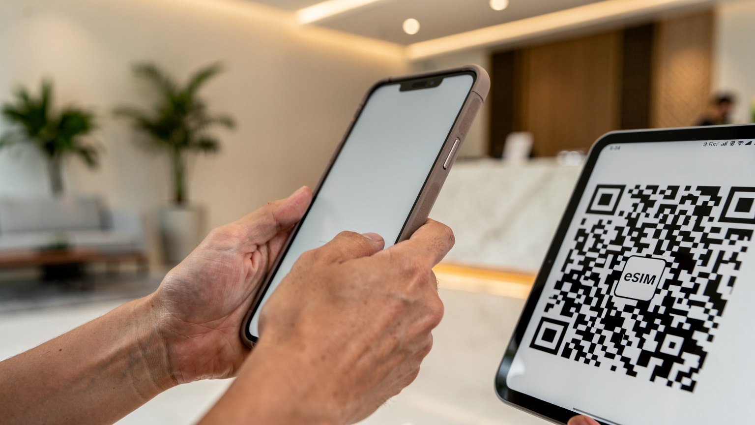 A person's hands holding an iPhone, scanning a QR code from another device's screen, with a blurred background of a modern hotel lobby in Singapore.
