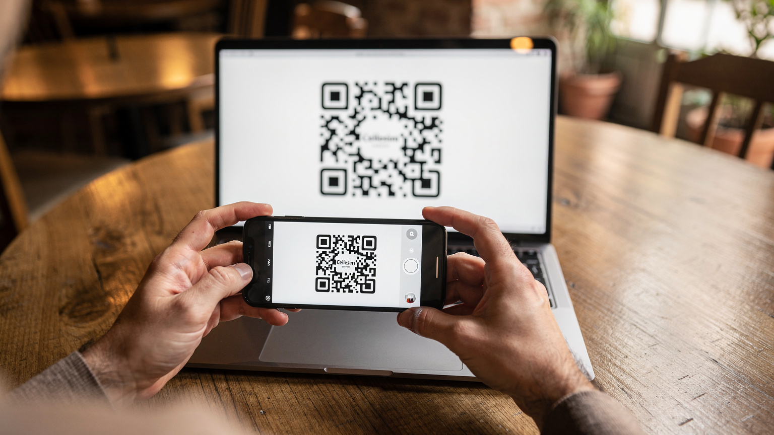 Човек сканира QR код на екрана на своя лаптоп с телефона си, за да активира eSIM, докато седи в уютно кафене в Барселона.