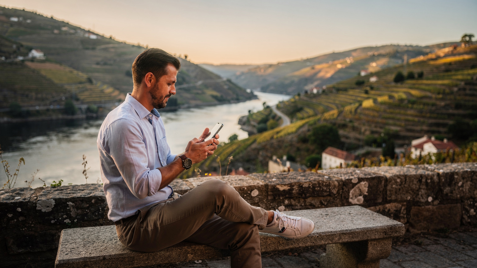 Um viajante a consultar o mapa de cobertura no seu smartphone, sentado num banco com vista para o rio Douro e as suas vinhas em socalcos, aproveitando o clima ameno de setembro.