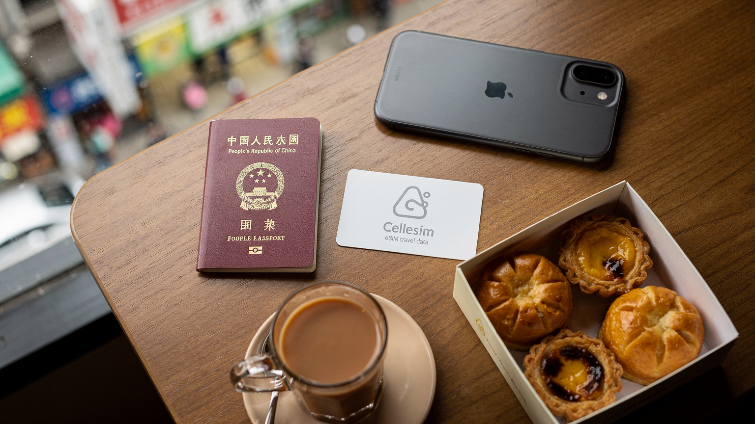 一张桌面平铺照片,展示了旅行者的必需品:一本中国护照、一张Cellesim的eSIM说明卡、一部智能手机、一盒印着香港特色美食(如菠萝包或蛋挞)的零食和一杯港式奶茶,背景是模糊的香港街景。