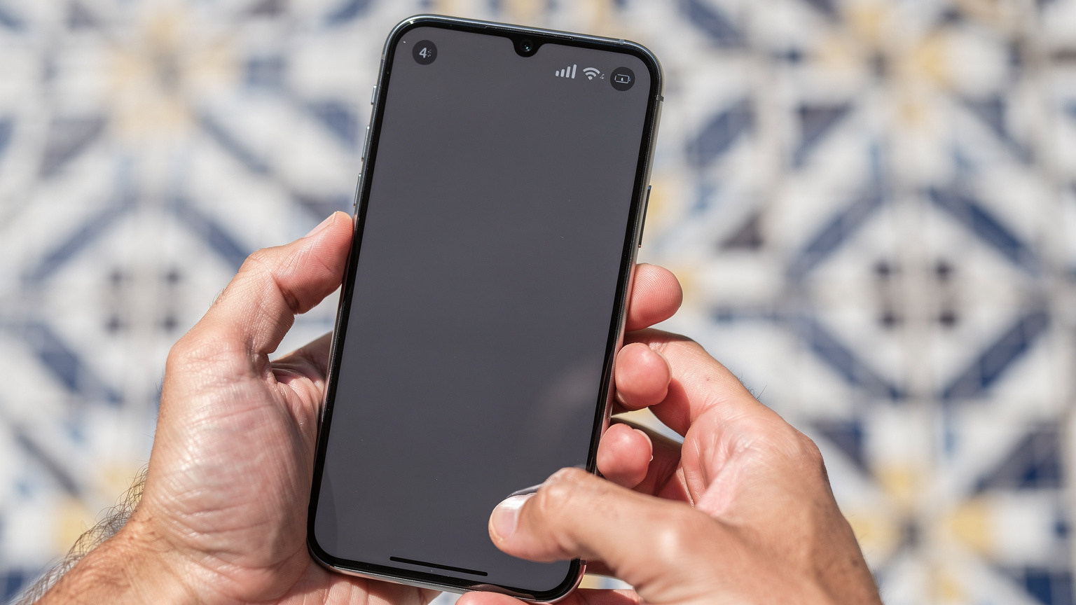 Zbliżenie na dłonie trzymające nowoczesny smartfon, z wyraźnie widoczną ikoną zasięgu sieci komórkowej oraz włączonym WiFi, na tle rozmytej, kolorowej mozaiki na portugalskiej ulicy, w słonecznym świetle.