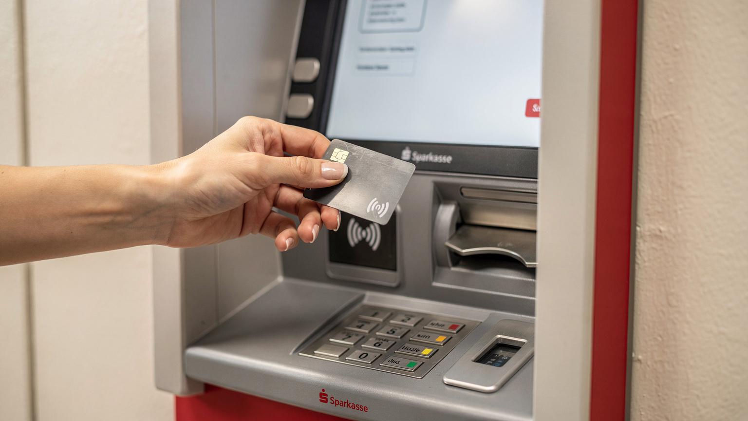 Primer plano de una mano sosteniendo una tarjeta de débito, interactuando con la pantalla táctil de un cajero automático de Sparkasse en Alemania, con el teclado numérico visible y sin signos de manipulación.