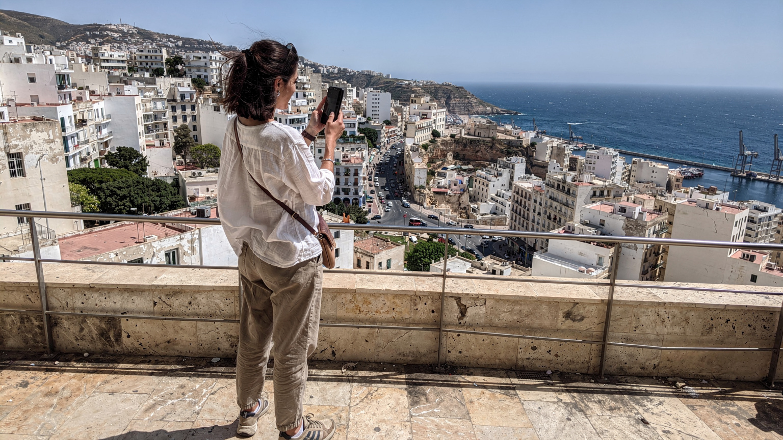 Une femme vérifie la couverture réseau sur son smartphone en haut d'une colline surplombant Alger, avec la ville s'étendant à l'horizon.