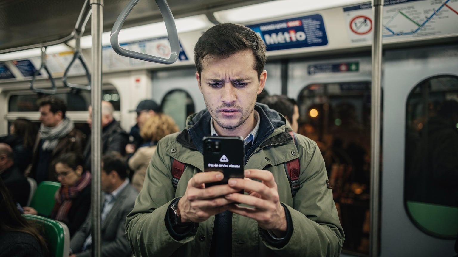 Un viajero con una expresión de preocupación, sosteniendo su teléfono y mirando una pantalla con un mensaje de error o sin señal, en medio de la multitud en el metro de París durante la hora punta.