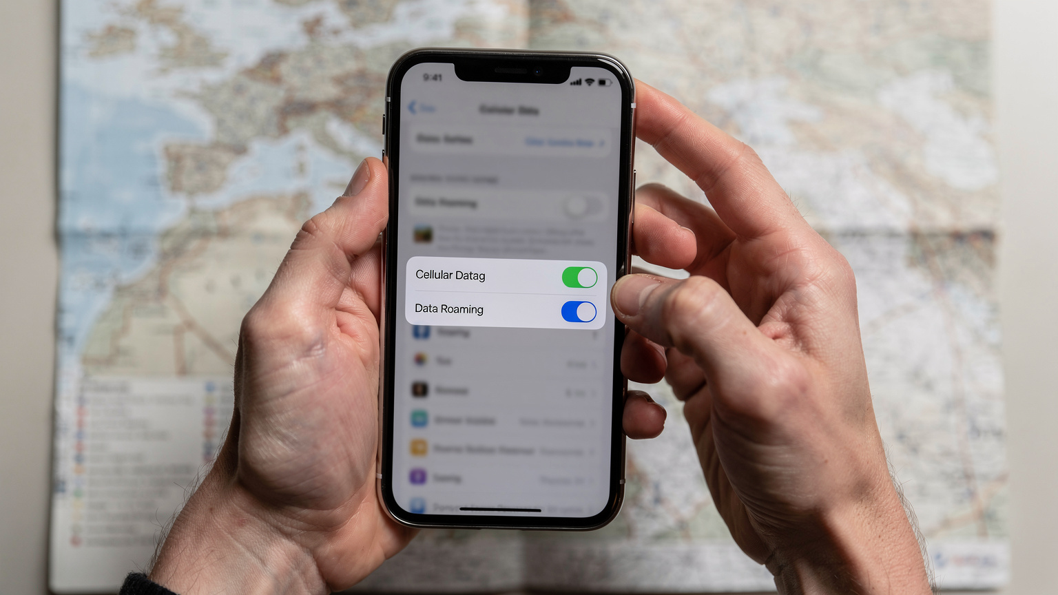 Gros plan sur des mains tenant un smartphone montrant la page des paramètres de données cellulaires, avec l'option "Données en itinérance" clairement désactivée pour la ligne principale, sur un fond flou de carte de transport ou de guide de voyage.
