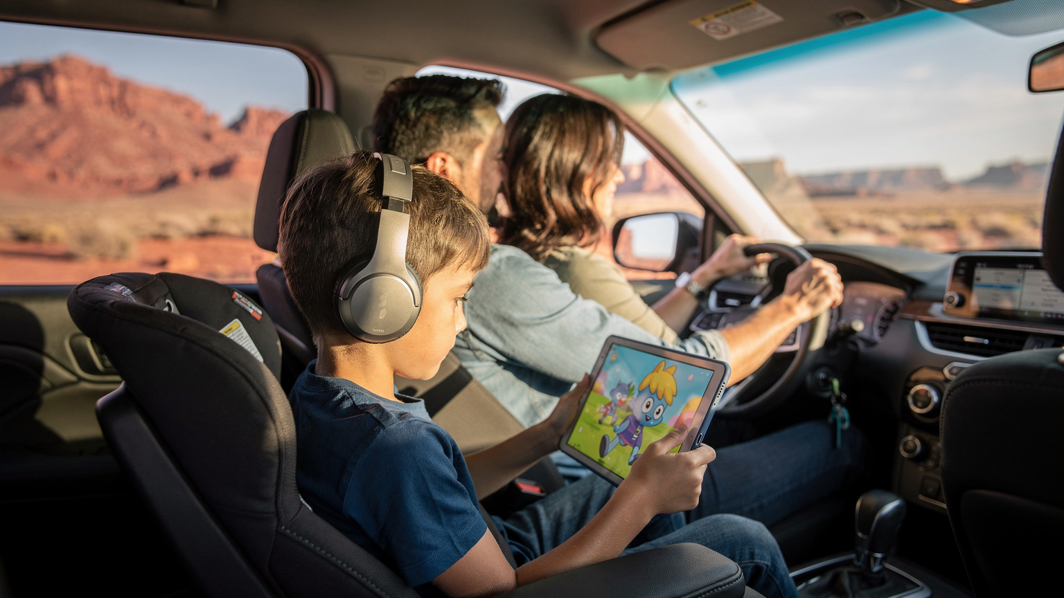 Un bambino con cuffie guarda un cartone animato sul tablet seduto sul sedile posteriore di un'auto, mentre i suoi genitori sono nel sedile anteriore e guidano attraverso un paesaggio desertico americano.