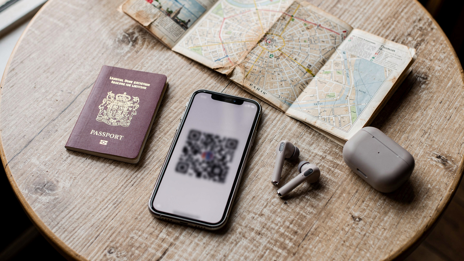 An overhead flat-lay shot of a traveler's essential items: a passport, a smartphone displaying a QR code, a pair of wireless headphones, and a small, folded city map of Paris, all arranged on a light wooden table.