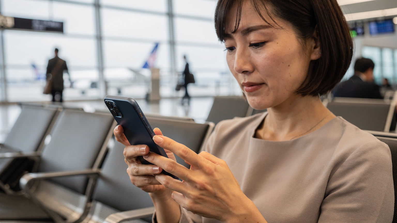 成田空港の搭乗口近くのベンチで、出発前の最終確認としてiPhoneの設定画面を真剣に見つめる母親のクローズアップ。
