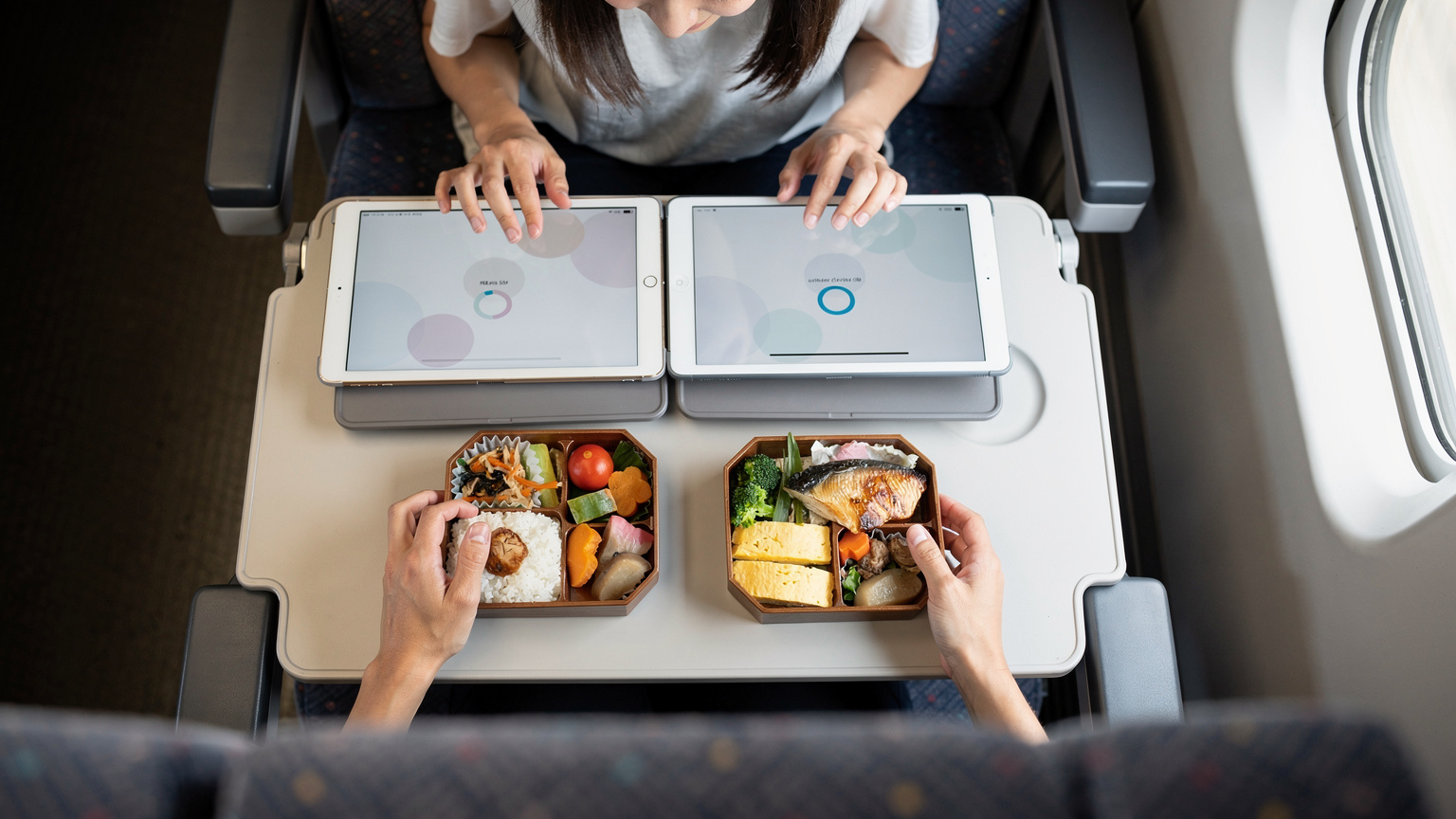 新幹線の中で、日本の駅弁が広げられたテーブルの上で、母親が子供たちのiPadとNintendo Switchにオフラインコンテンツをダウンロードしているオーバーヘッドフラットレイ。