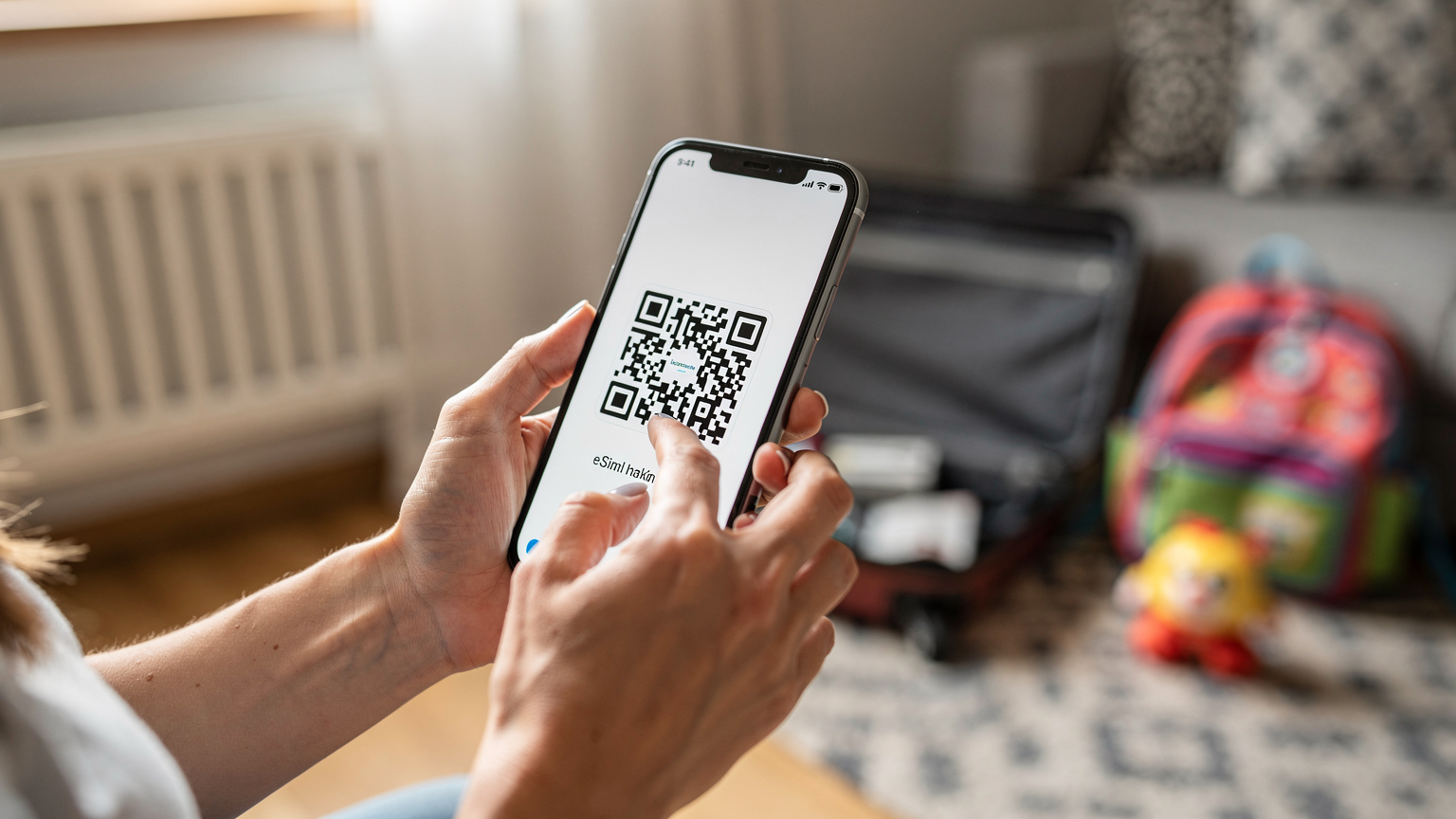 Zbliżenie na dłonie matki trzymającej smartfon, na ekranie którego widać fragment kodu QR, a w tle rozmyte walizki i dziecięce zabawki, sugerujące przygotowania do podróży.
