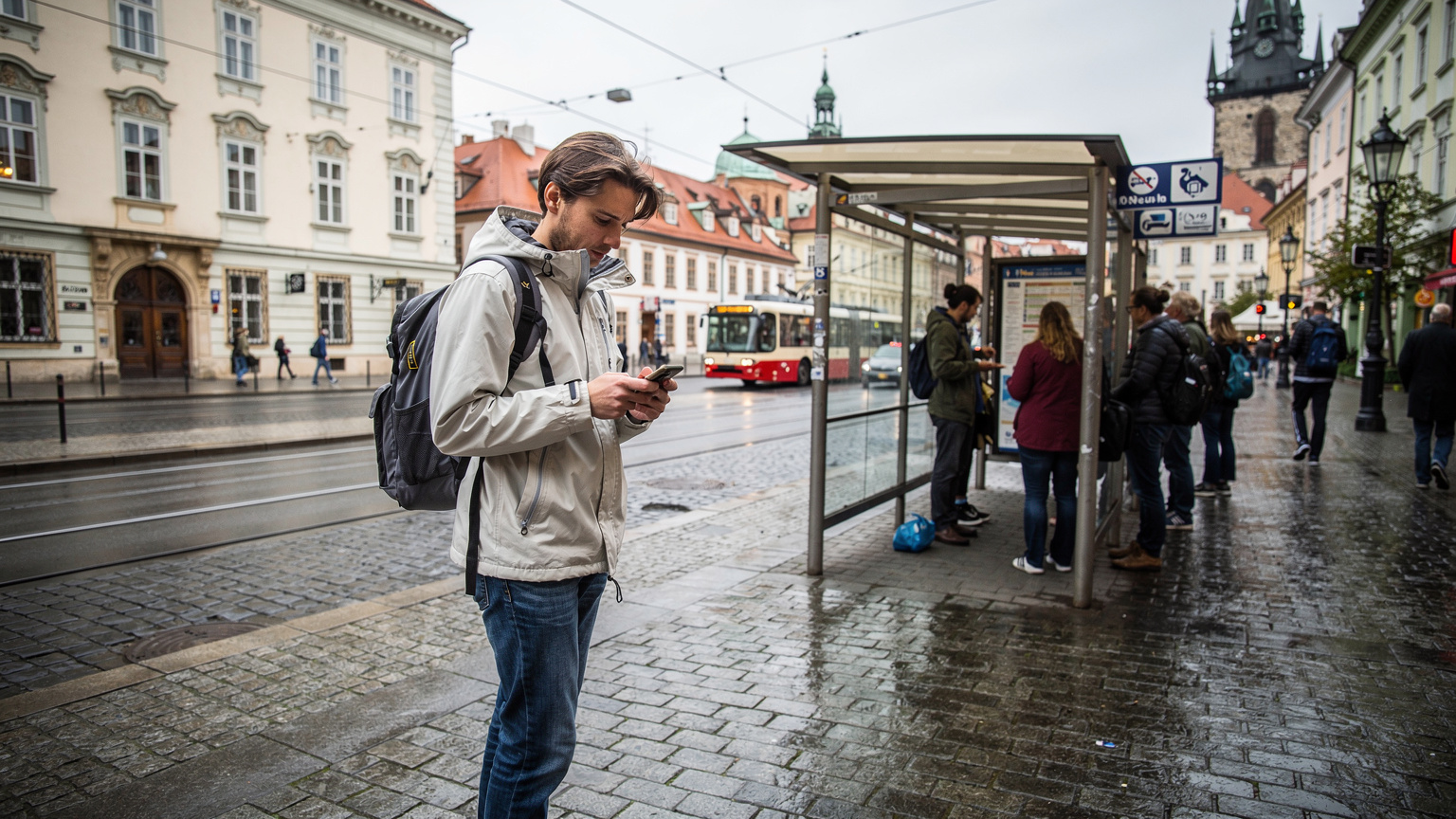 En bild på en europeisk gatubild med en tågstation eller busshållplats i förgrunden, där en resenär tittar på sin telefon med en fundersam min, kanske över täckningsproblem.