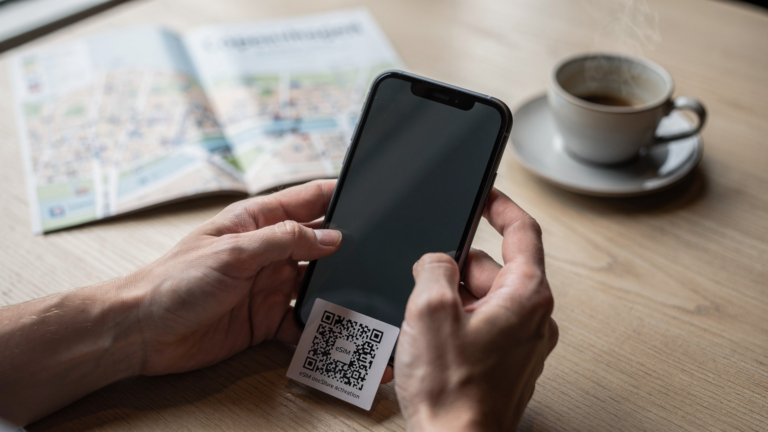 En detaljbild av en resenärs händer som håller en smartphone och är på väg att skanna en QR-kod för eSIM-aktivering, med en resguide eller karta skymtande i bakgrunden.