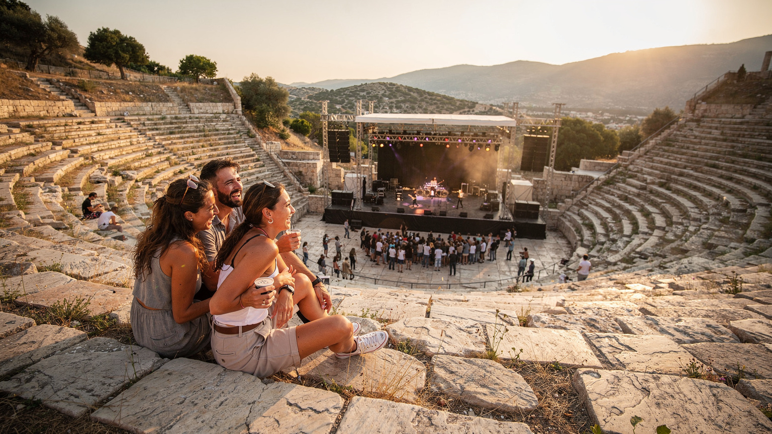 Μια ομάδα φίλων χαμογελά ενώ παρακολουθεί μια συναυλία σε ένα αρχαίο ελληνικό θέατρο, με το ηλιοβασίλεμα να φωτίζει τη σκηνή.