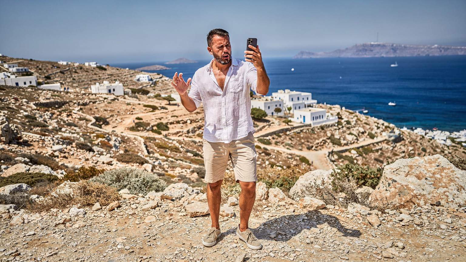 Ένας άντρας κοιτάζει το smartphone του με απογοήτευση, προσπαθώντας να βρει σήμα σε ένα απομακρυσμένο ελληνικό νησί, με φόντο έναν ξηρό λόφο και τη θάλασσα.