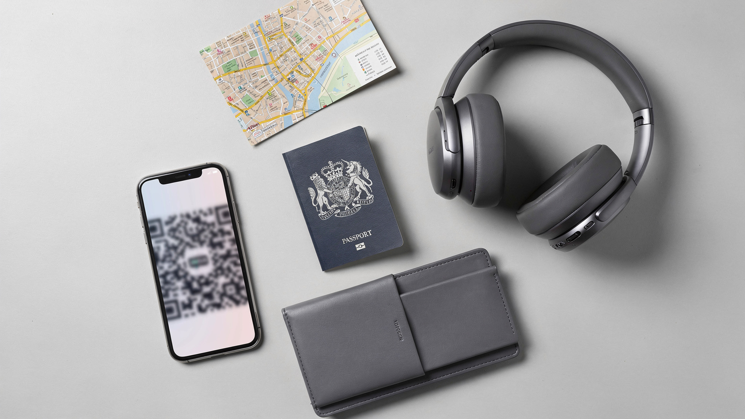 An overhead flat-lay of travel essentials: a passport, a smartphone with an eSIM QR code partially visible, noise-cancelling headphones, a small physical map of a European city, and a travel wallet, all neatly arranged on a light-coloured surface.