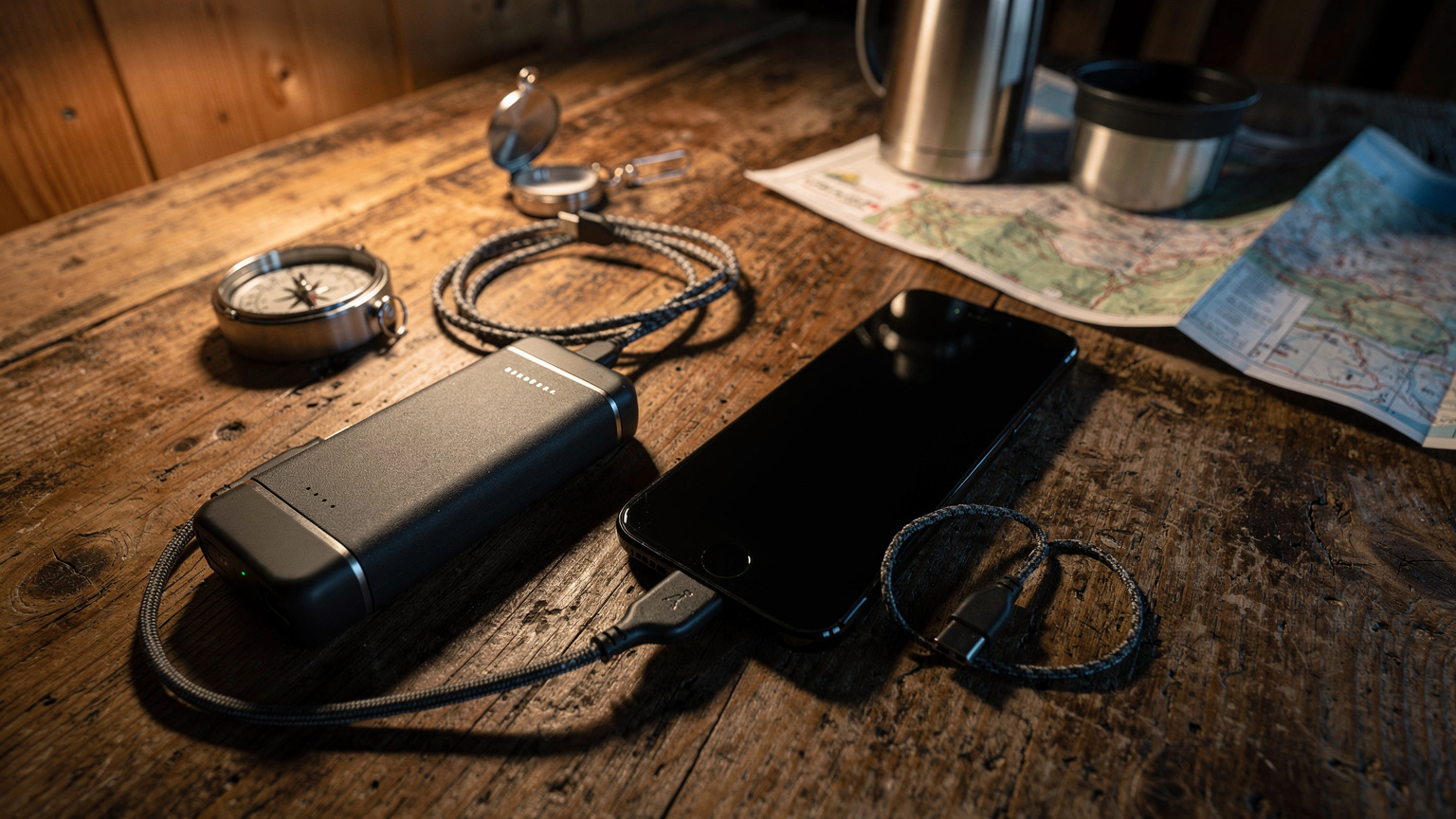 Eine Nahaufnahme eines Smartphones, das neben einem Powerbank und einem Ladekabel auf einem hölzernen Tisch in einer Berghütte liegt, im Hintergrund verschwommen Wanderutensilien.