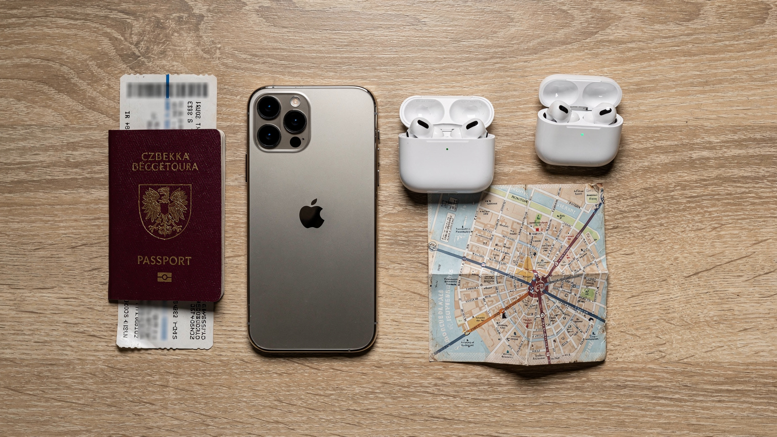 Cestovní flat-lay: iPhone 18 Pro Max, pas České republiky, letenky, bezdrátová sluchátka a mapa Paříže ležící na dřevěném stole, připravené k odjezdu.