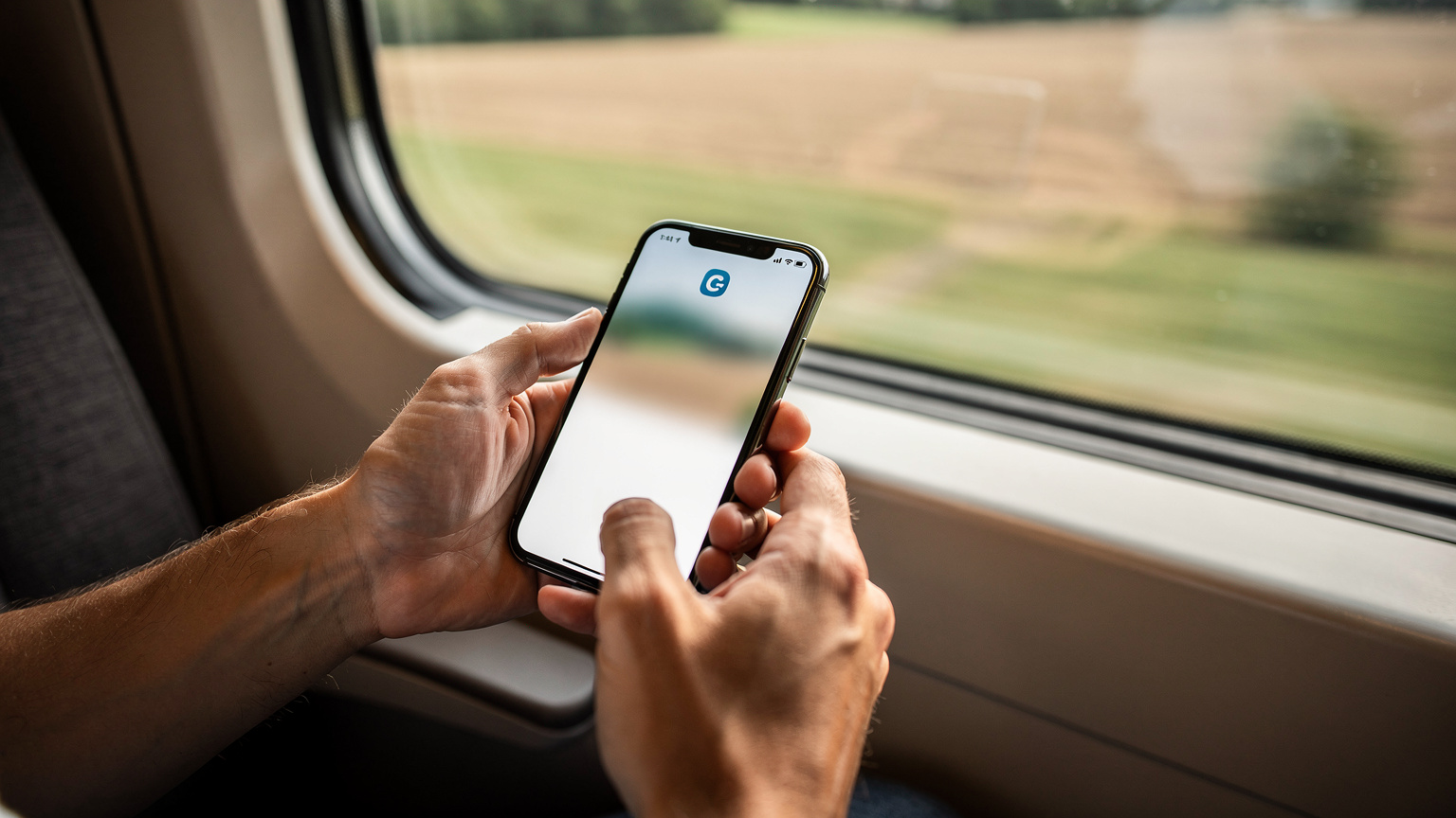 ヨーロッパを列車で移動中にスマートフォンでCellesimアプリからデータリチャージをしている旅行者の手元。車窓には美しい田園風景が広がっている。