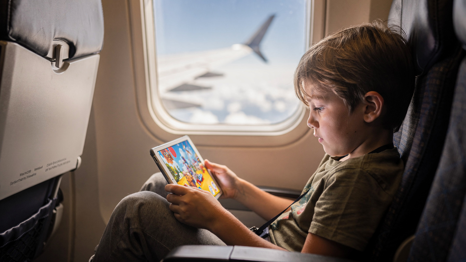 Ein Kind im Alter von etwa 7 Jahren schaut fasziniert auf ein Tablet, auf dem ein Disney+-Video läuft, während es entspannt in einem Flugzeugsitz sitzt. Im Hintergrund ist ein Teil des Flügels und die Wolken zu sehen.