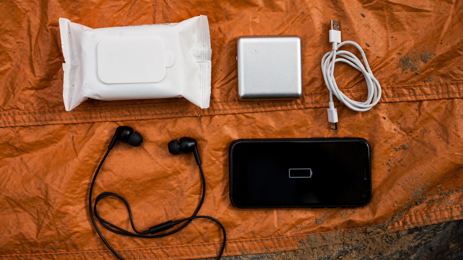 Et editorial overhead flat-lay af en pakke vådservietter, en powerbank, et par in-ear hovedtelefoner, og en smartphone, der viser et lavt batteriniveau, arrangeret på en teltdug, der indikerer festivalens udfordringer.
