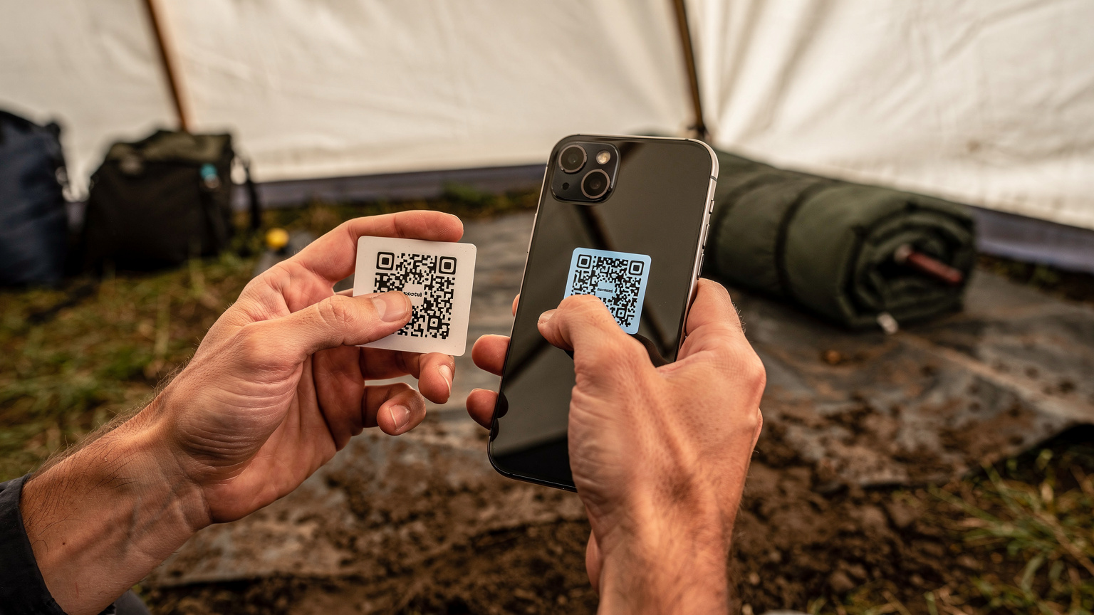 En tæt billed af hænder, der holder en smartphone og scanner en QR-kode for at aktivere et eSIM, med en baggrund af et telt og festivaludstyr, der antyder en hurtig og nem opsætning i en camping kontekst.