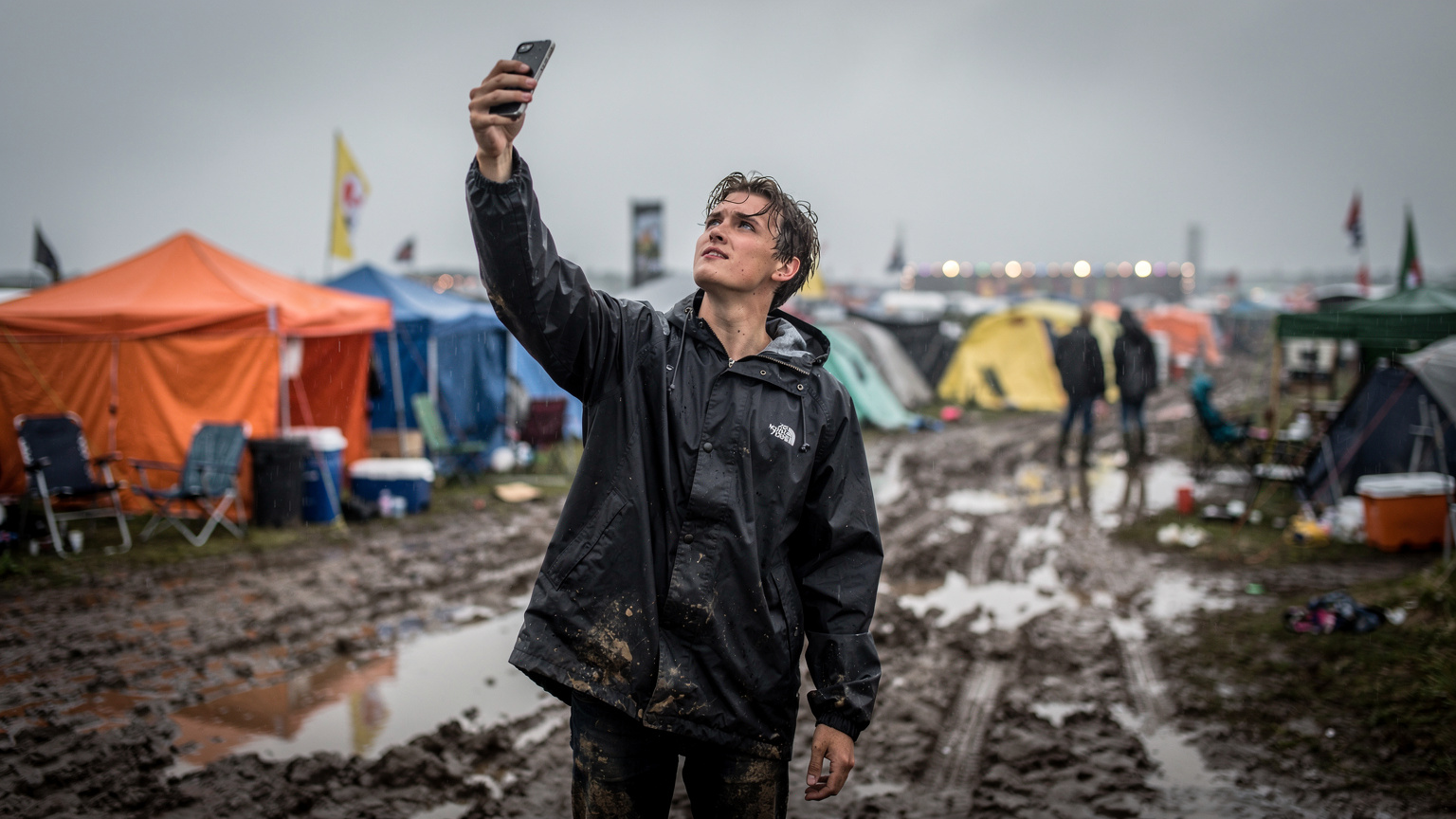 En ung mand i regnjakke holder sin smartphone op i luften, idet han forsøger at finde mobilsignal på en våd festivalplads, med telte og mudder i forgrunden og en overskyet himmel.