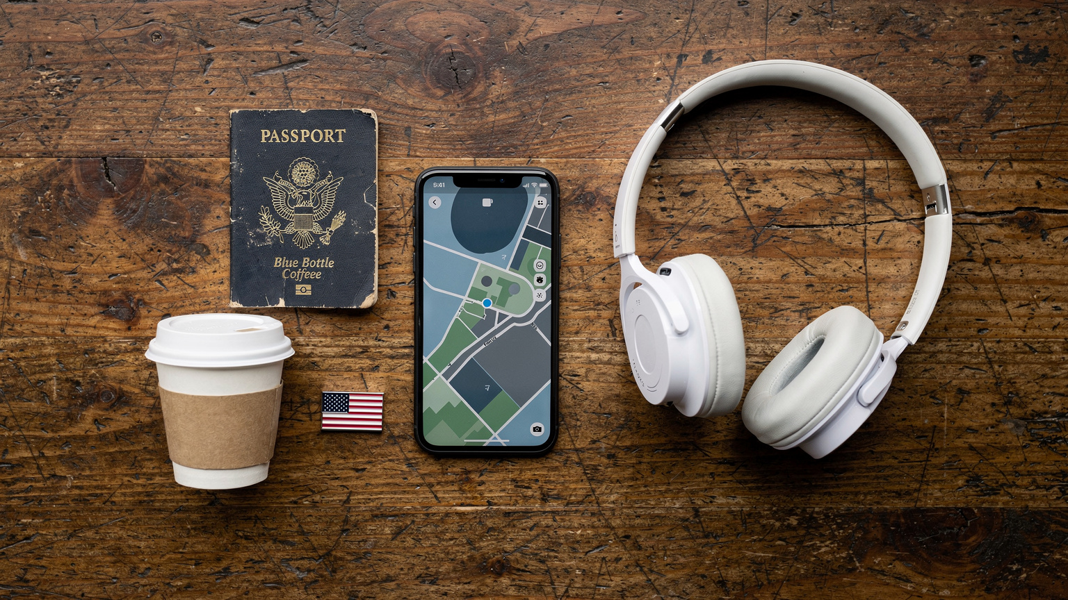 An overhead flat-lay of essential travel items for a USA trip: a passport, a smartphone showing a map application, wireless headphones, a coffee cup, and a small US flag pin, on a rustic wooden table.