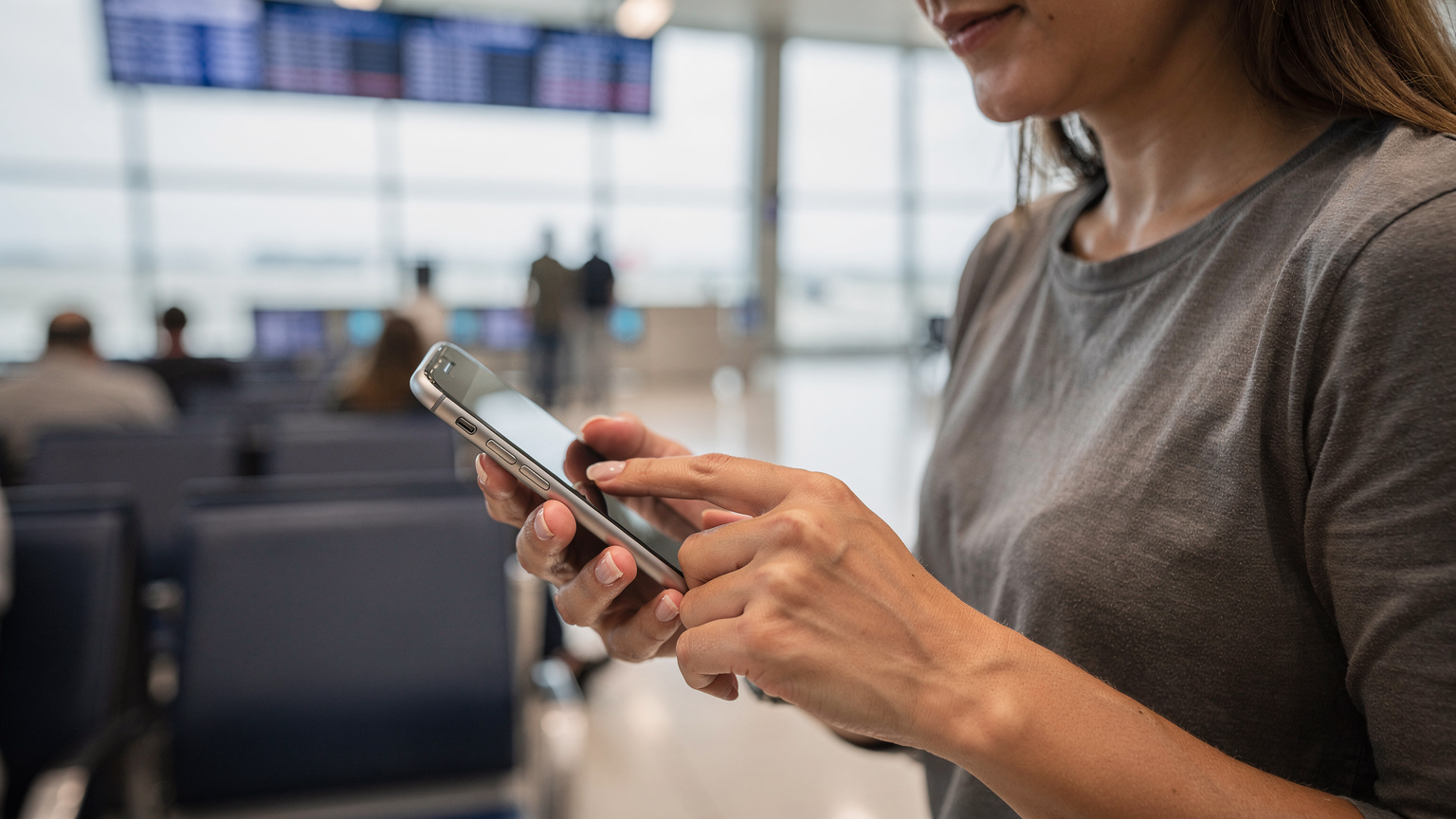 Cestovatelka se soustředěně dotýká obrazovky smartphonu, na kterém je vidět rozhraní nastavení eSIM. Její ruce jsou viditelné, ale žádný text na obrazovce není čitelný. V pozadí je rozmazané letištní prostředí.