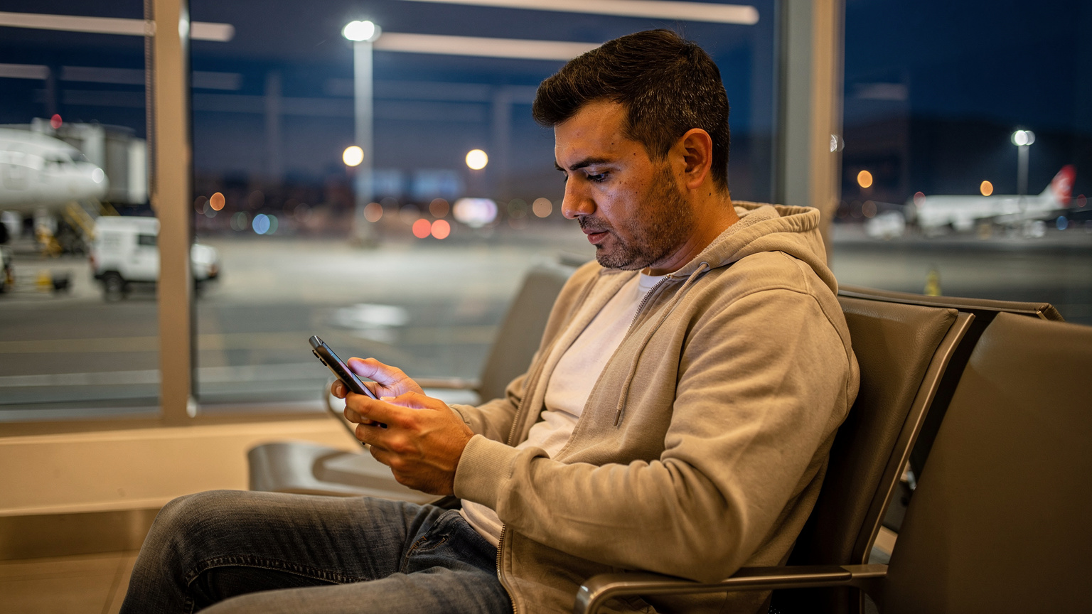 Un viajero en el Aeropuerto Internacional Jorge Chávez en Lima, Perú, activa su eSIM desde su teléfono, con las luces de la pista de aterrizaje visibles a través de una ventana borrosa.
