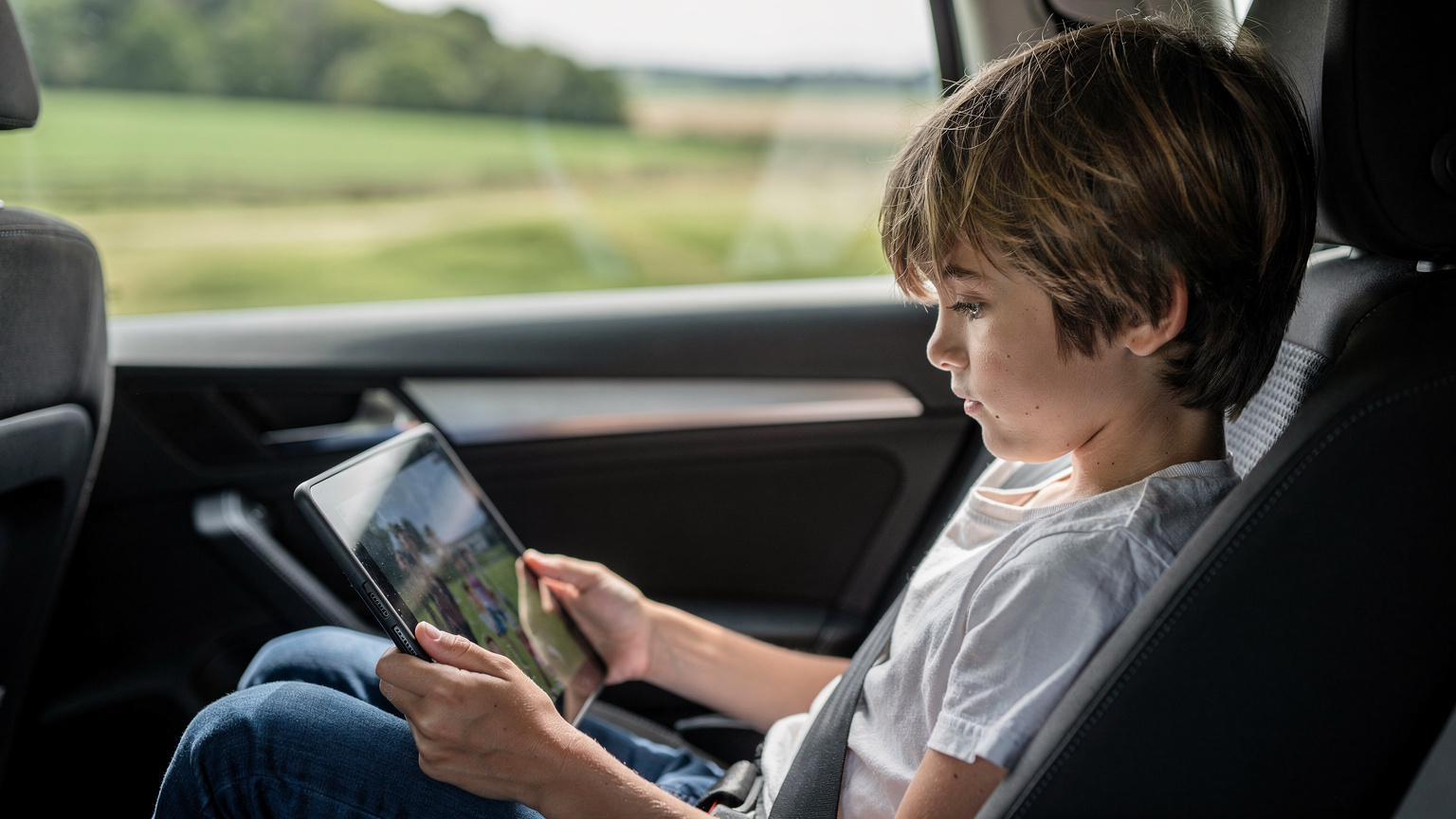 Ein Kind auf dem Rücksitz eines Autos schaut entspannt auf ein Tablet, das Videos streamt, während die Landschaft draußen vorbeizieht.