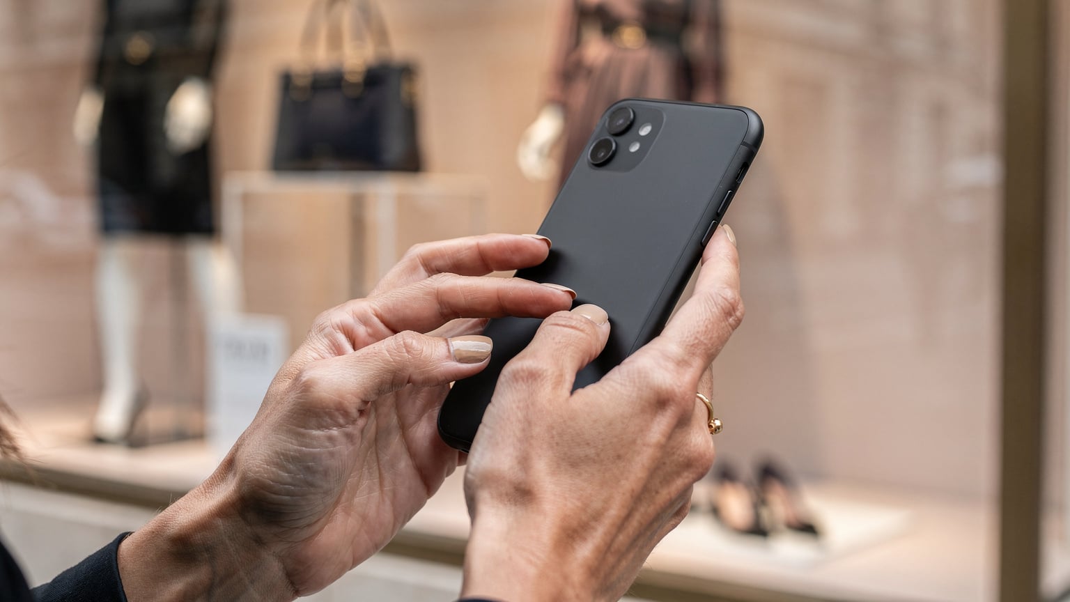 Un primo piano delle mani di una donna italiana mentre confronta prezzi e recensioni di prodotti di moda sul suo smartphone, con lo sfondo sfocato di una vetrina di un negozio di lusso a Boulevard Haussmann, Parigi.