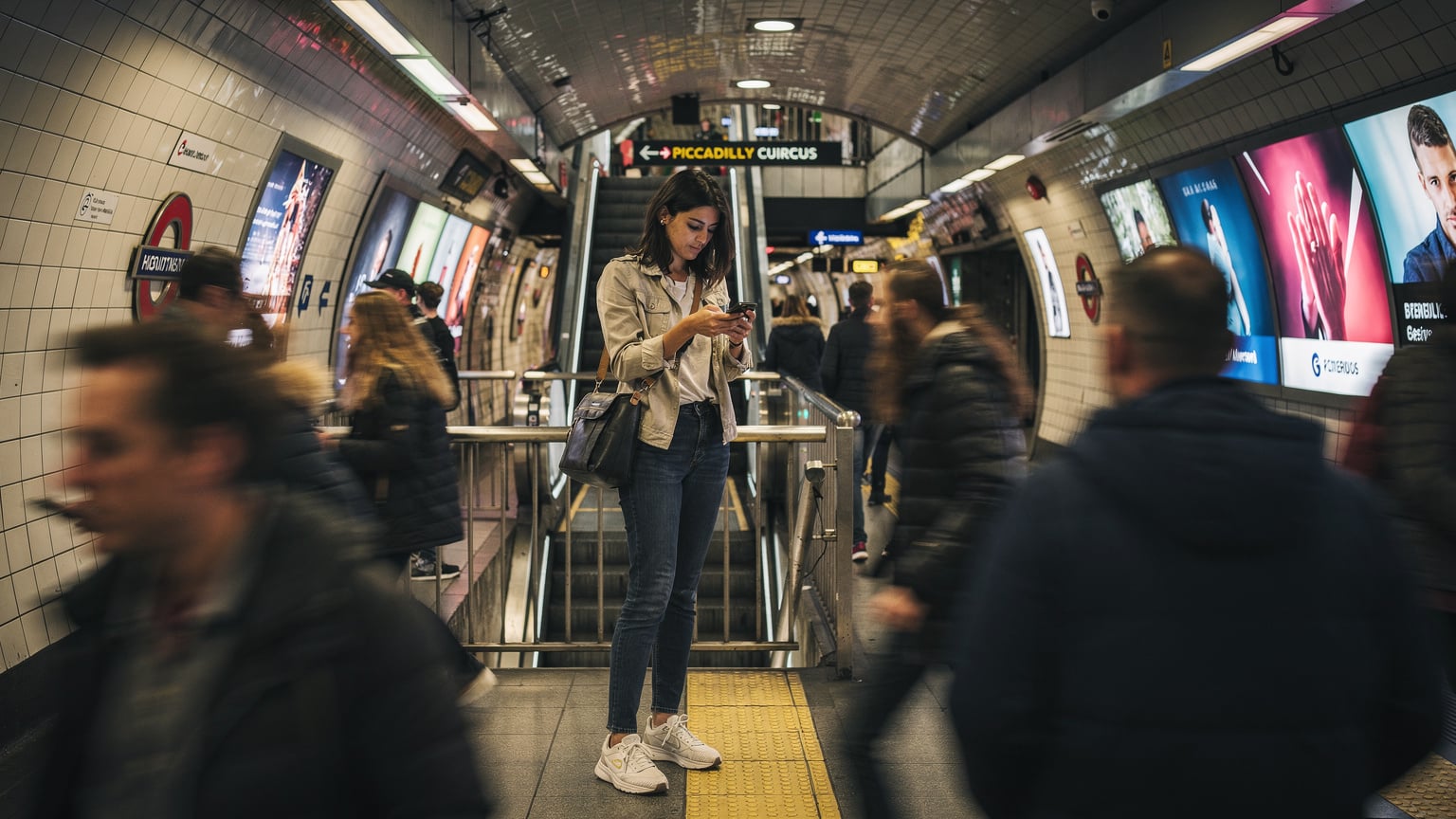 Una viaggiatrice italiana consulta il suo smartphone per trovare la fermata della metropolitana più vicina, immersa nel flusso di persone all'interno della stazione di Piccadilly Circus a Londra, tra luci al neon e cartelloni pubblicitari.