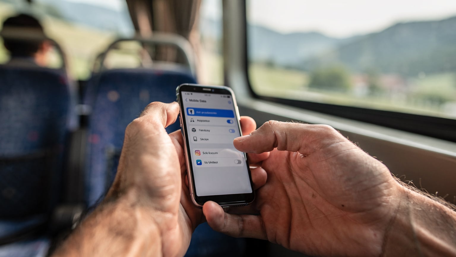 Detailní záběr rukou cestovatele držícího smartphone s otevřeným nastavením mobilních dat, kde je vidět aktivní eSIM profil pro Albánii a Černou Horu, s rozmazaným pozadím interiéru autobusu na cestě.