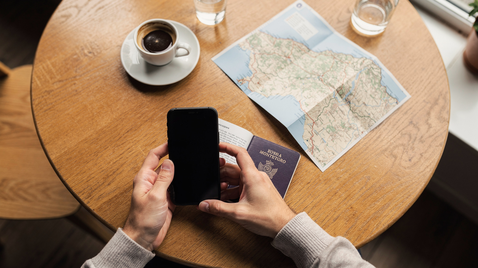 Zblízka pohled na ruce držící mobilní telefon a cestovní pas na dřevěném stole v kavárně v Podgorici, s šálkem turecké kávy a mapou Černé Hory v pozadí, s měkkým ranním světlem.