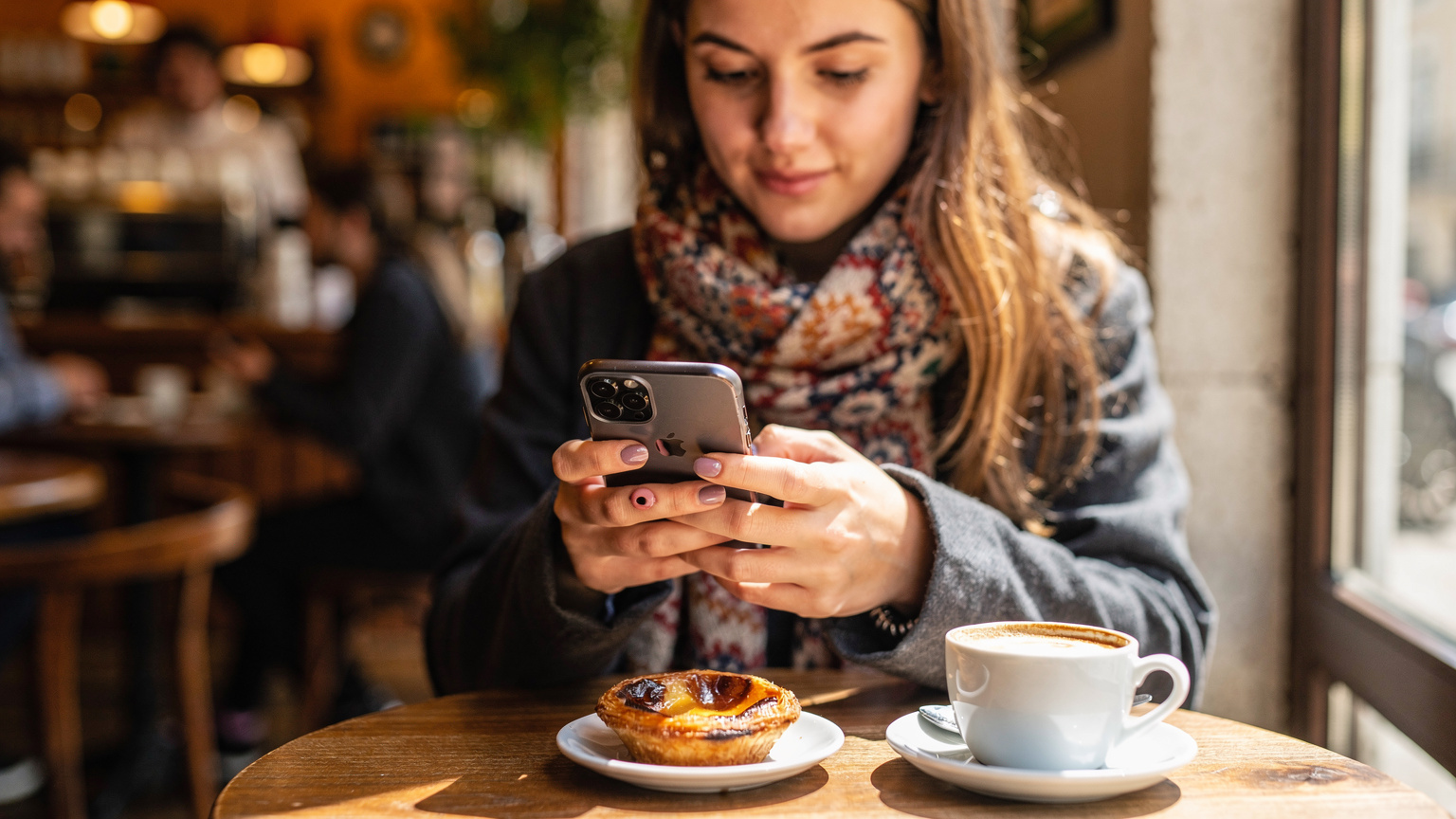 Un călător verifică compatibilitatea eSIM pe smartphone-ul său într-o cafenea aglomerată din Lisabona, cu o cafea și o patiserie tipic portugheză pe masă.
