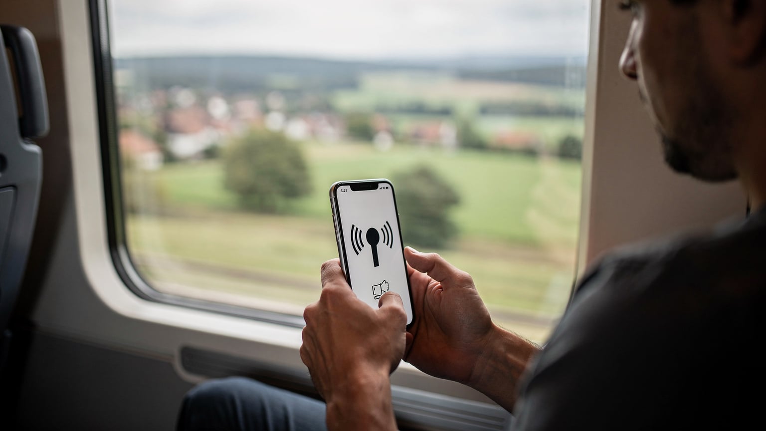 O persoană într-un tren de mare viteză european, privind pe fereastră la peisajul rural care trece, cu un smartphone în mână afișând un ecran de "fără semnal" sau "căutare rețea".