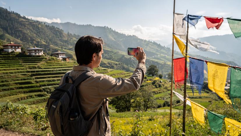 A traveler checks their phone for signal strength in a rural Bhutanese valley, with traditional prayer flags fluttering in the background, highlighting the contrast between ancient traditions and modern technology.