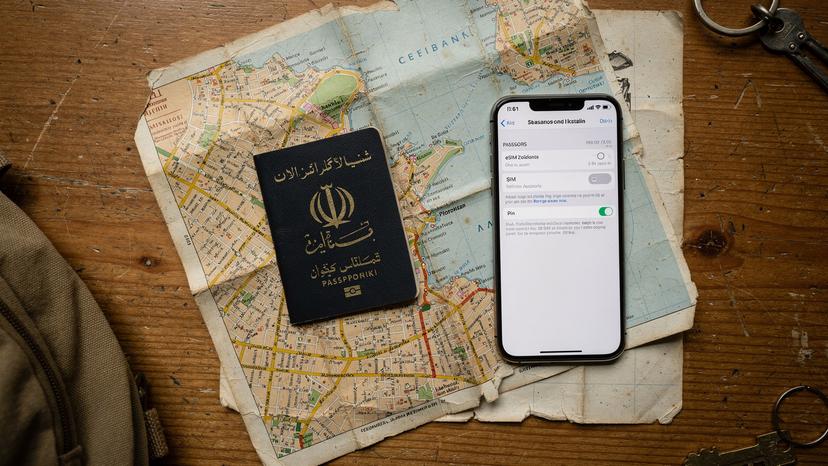 یک پاسپورت ایرانی، یک گوشی هوشمند با صفحه تنظیمات eSIM و یک نقشه کاغذی قدیمی از سالونیک، یونان، روی میز یک هاستل ارزان.