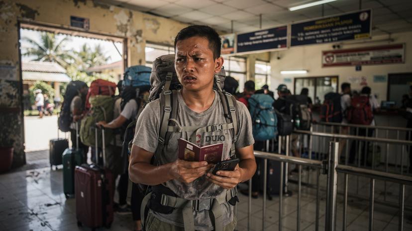 A solo backpacker looking frustrated, holding their phone and an open passport, standing in a long, confusing queue at a land border crossing in Southeast Asia, with signs in multiple languages.