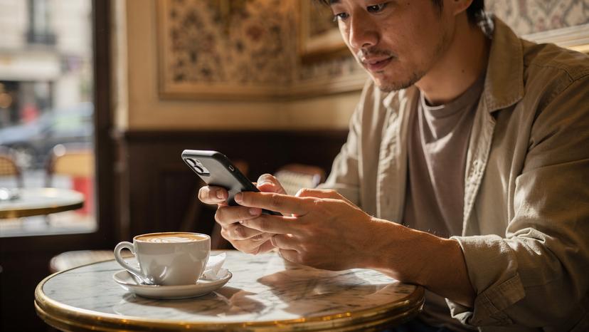 パリのカフェで、ラテを飲みながらスマートフォンでeSIMに関するよくある質問を調べている日本人男性旅行者。旅の準備を抜かりなく進める様子。