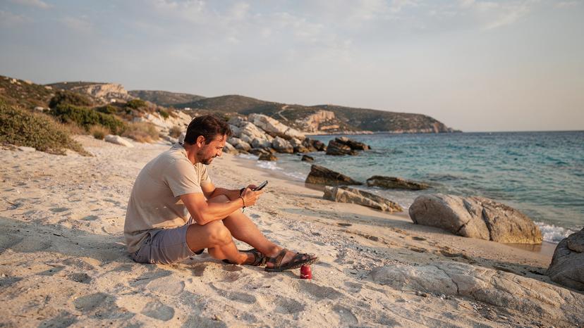 En ensam resenär sitter på en avlägsen strand på en grekisk ö, tittar på sin smartphone med en svag signalindikator, symboliserande utmaningar med täckning på avlägsna platser.
