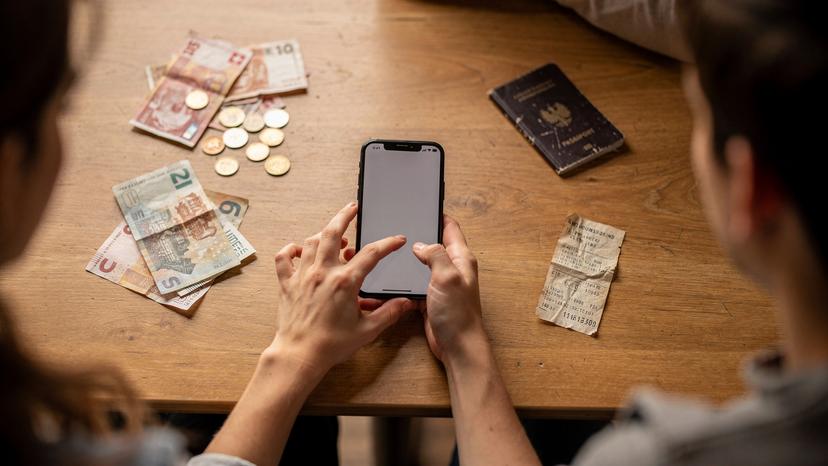 A female traveler's hands holding a smartphone, comparing various Cellesim eSIM plans for Switzerland, with a scatter of Swiss Franc notes and coins on a hostel common room table, emphasizing budgeting.