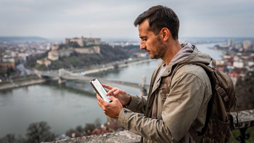 Un călător folosește o aplicație de hărți pe telefon, având în fundal o imagine panoramică a orașului Belgrad, cu fortăreața Kalemegdan și râurile Sava și Dunărea.