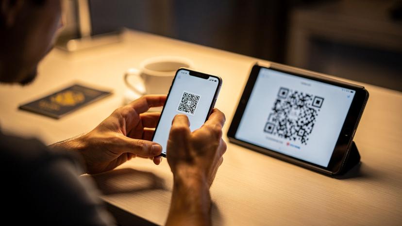 Un primer plano de manos de un viajero utilizando su smartphone para escanear un código QR desde otro dispositivo, con una taza de café y un pasaporte al fondo en un escritorio de hotel.