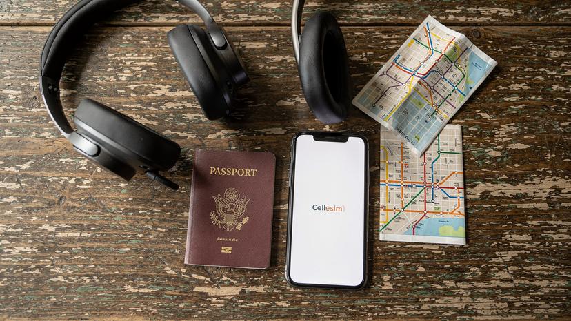 An overhead flat-lay shot of a traveler's essentials, including a passport, a smartphone showing a Cellesim logo on its screen (no readable UI), headphones, and a small, folded map, all resting on a rustic wooden table in a cafe, ready for a trip.