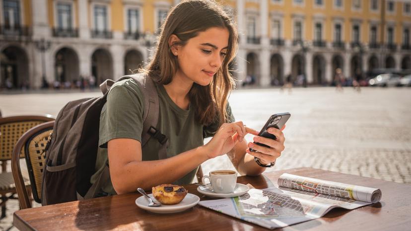 Woman in Lisbon cafe with iPhone 11, tapping screen, exploring eSIM options. Pastel de nata and espresso on table.