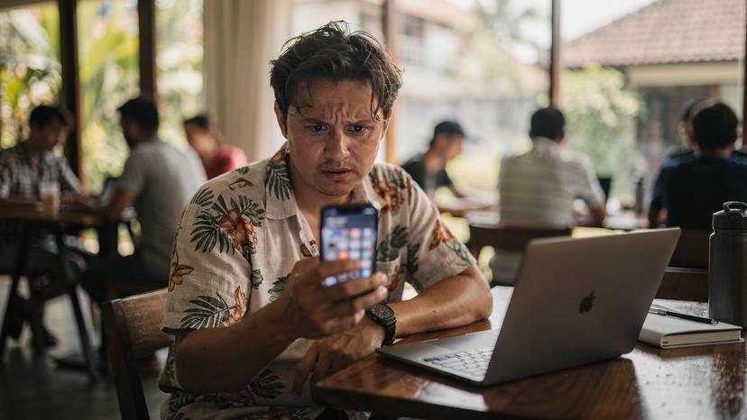 A digital nomad looking frustrated at their phone screen inside a bustling co-working space in Seminyak, Bali. The background shows other people working, with soft natural light filtering in from large windows. The phone screen is visible but blurred, conveying a sense of digital frustration.