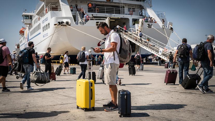 A digital nomad waiting at Piraeus port gate E7 with luggage and a smartphone in hand.